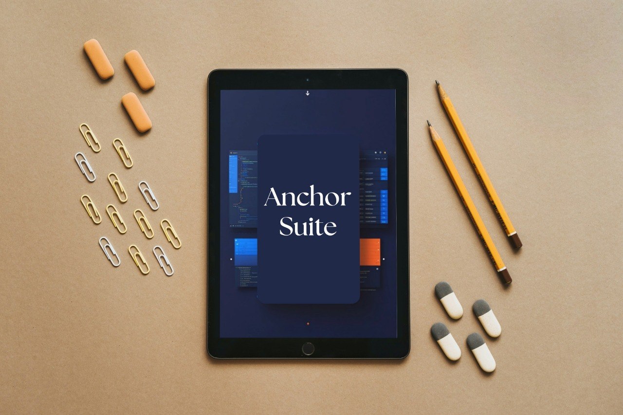 A tablet centered on a tan surface, displaying the text 'Anchor Suite' on its screen. Surrounding the tablet are orange earbuds, paperclips, yellow pencils, and white-and-gray capsules in a flat-lay arrangement.