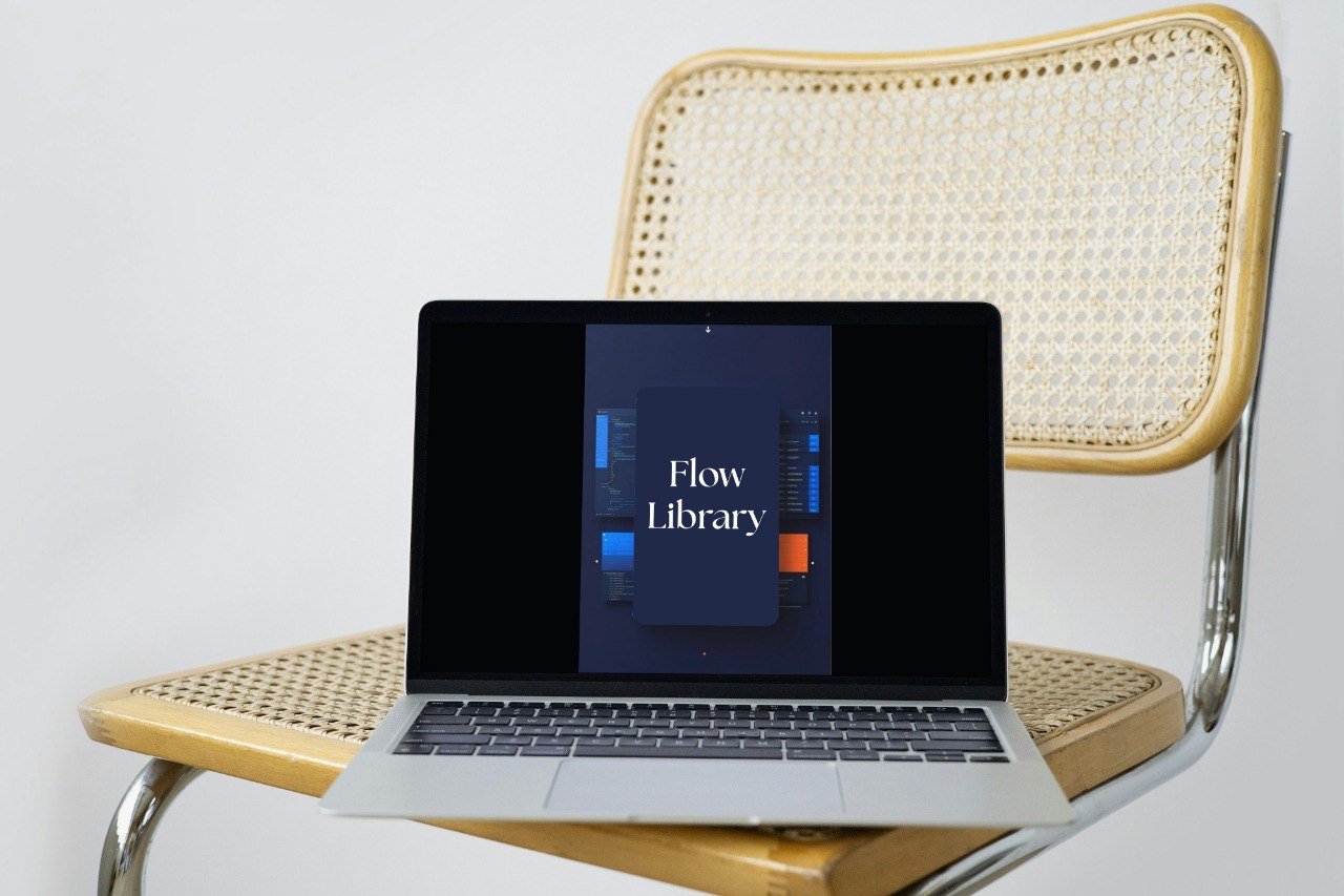 A silver laptop displaying the 'Flow Library' app on its screen, resting on a modern wooden and cane chair against a plain white background.